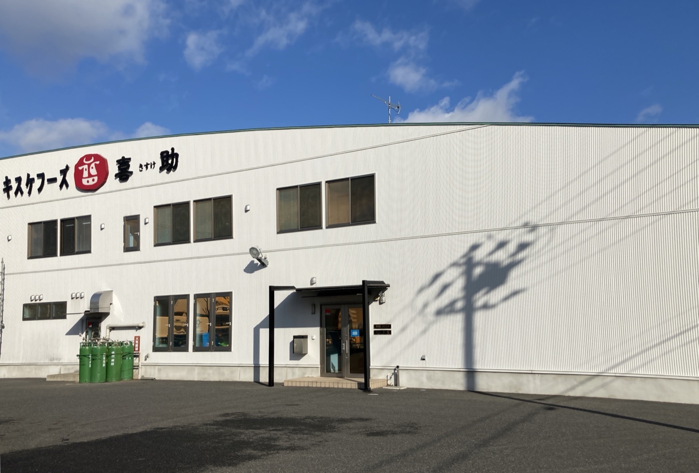 青空の下に建つキスケフーズ本社工場の外観。白い壁面に赤い喜助のロゴが掲げられている