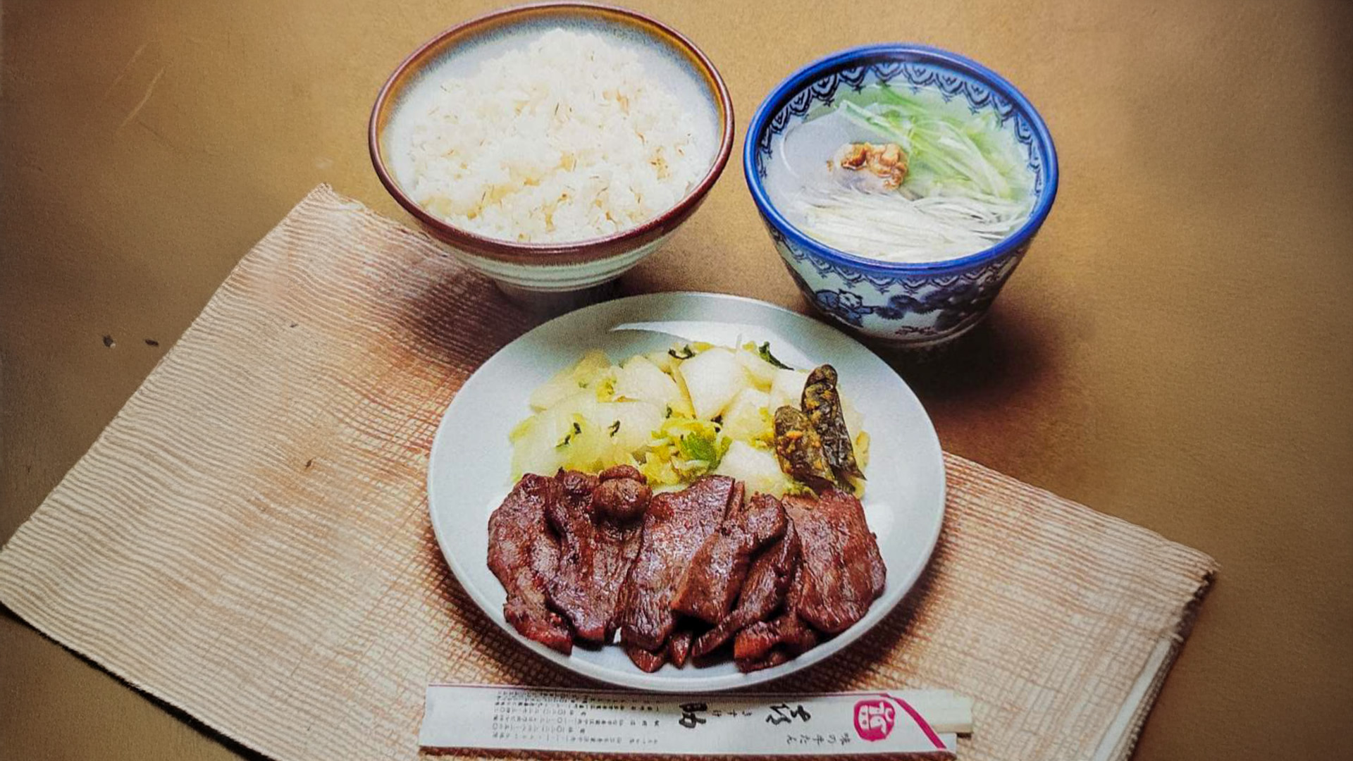 創業のころの牛たん定食。麦飯の茶碗、白髪ねぎを浮かべた染付の碗のテールスープ、牛たん焼きとお新香とみそ南蛮を盛った皿、喜助の箸袋が敷物の上に並んでいる